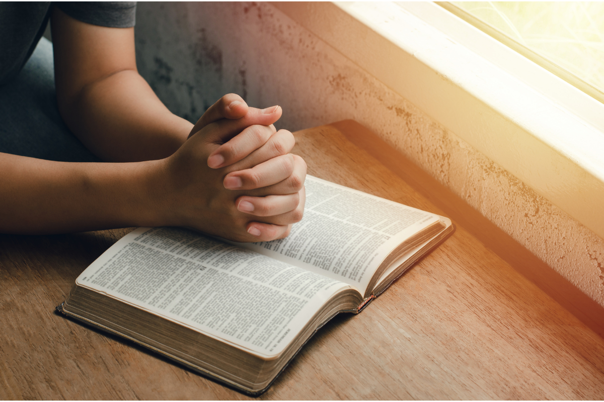 Hands folding in prayer over a Bible.