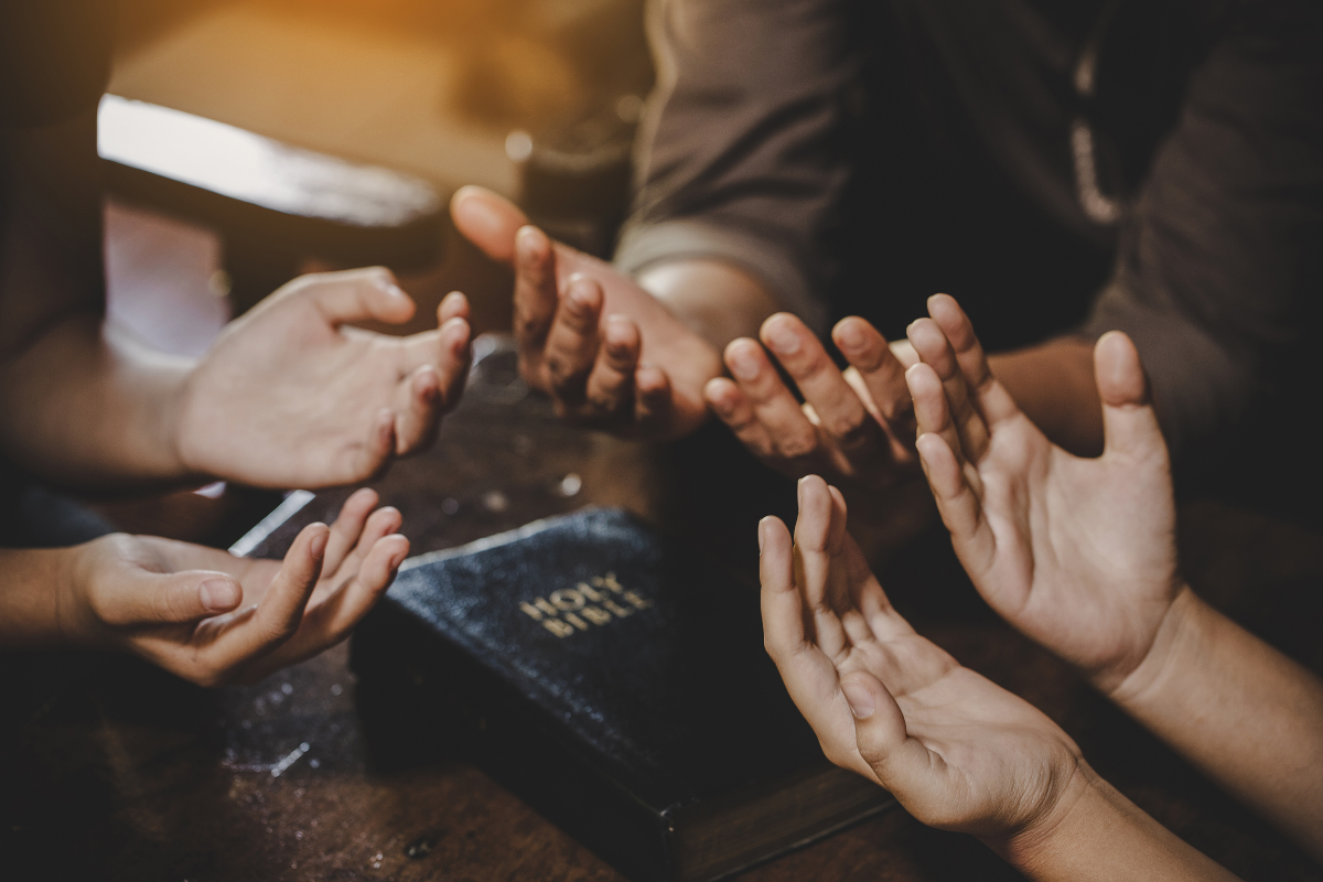 An image of hands in an act of praying with a Bible in between them.