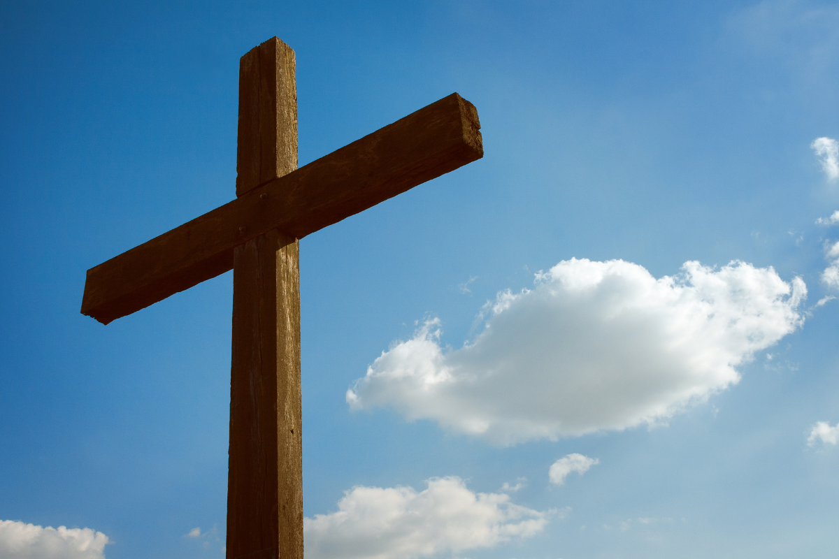 A wooden cross with a blue sky and clouds in the background.