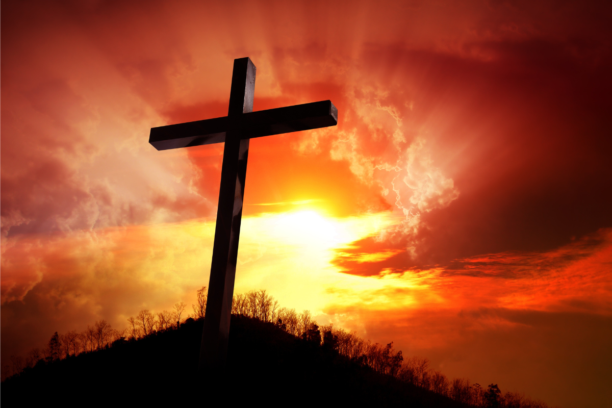 A silhouette of a cross on top of a hill, in a sunset background.