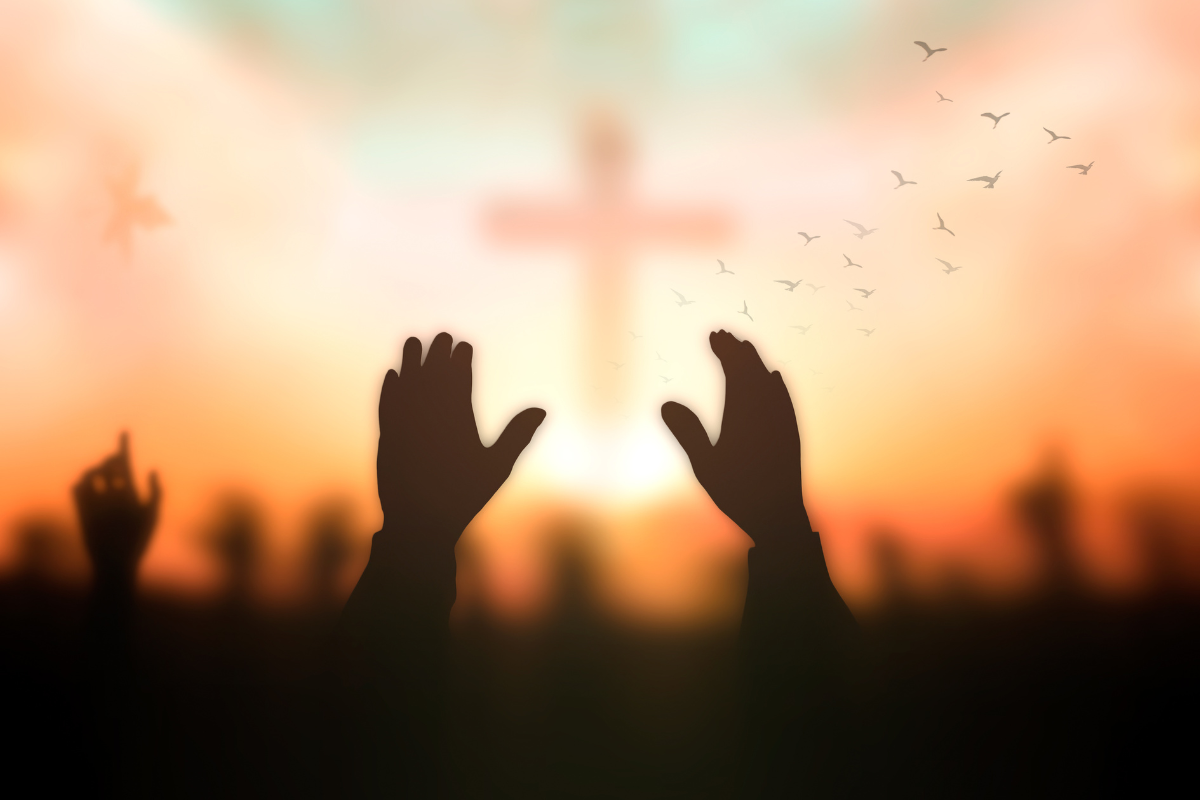 A pair of hands raised high, with a silhouette of a cross in the background.
