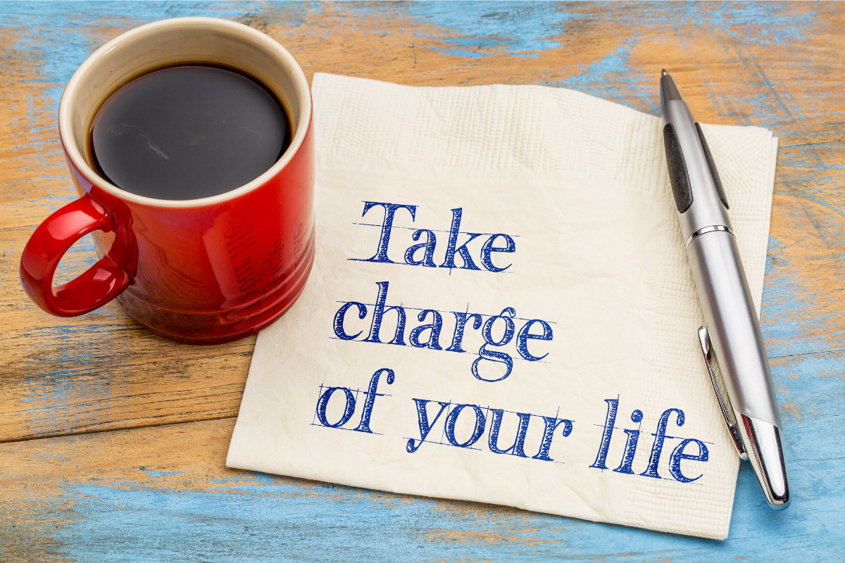 A red mug with coffee, a ballpoint pen, and a piece of paper with the phrase "Take charge of your life" written on it.