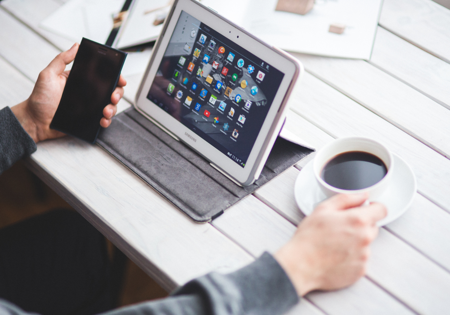 A person facing a tablet, while holding a phone and a coffee mug on the right.