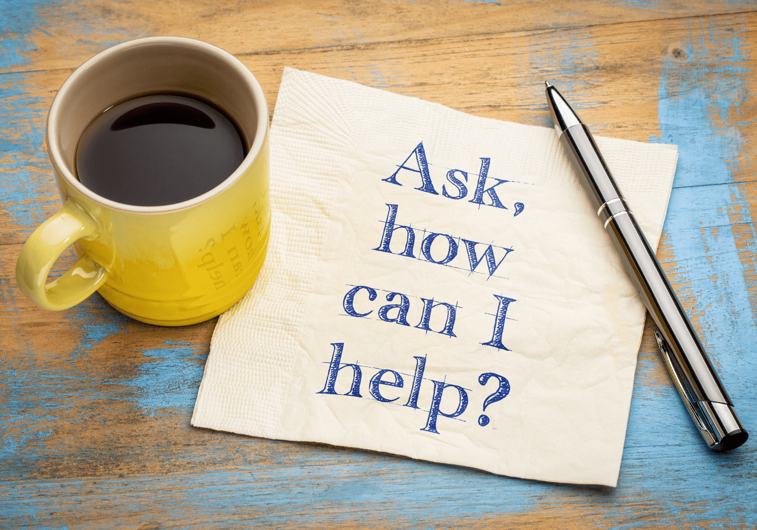 A yellow coffee mug with the words ASK, HOW CAN I HELP? written on a table napkin.